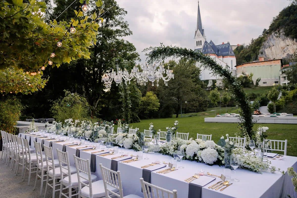 An elegant outdoor wedding reception setup with a long table adorned with white flowers and gold cutlery, set under a tree with string lights, with a picturesque view of a church in the background.