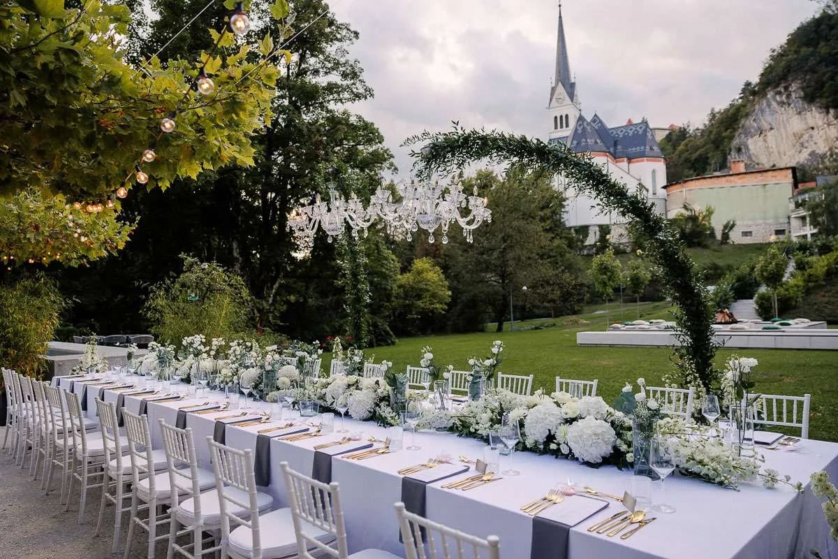 An elegantly set outdoor wedding reception table at Rose hotel adorned with white flowers, with a church and lush greenery in the background.