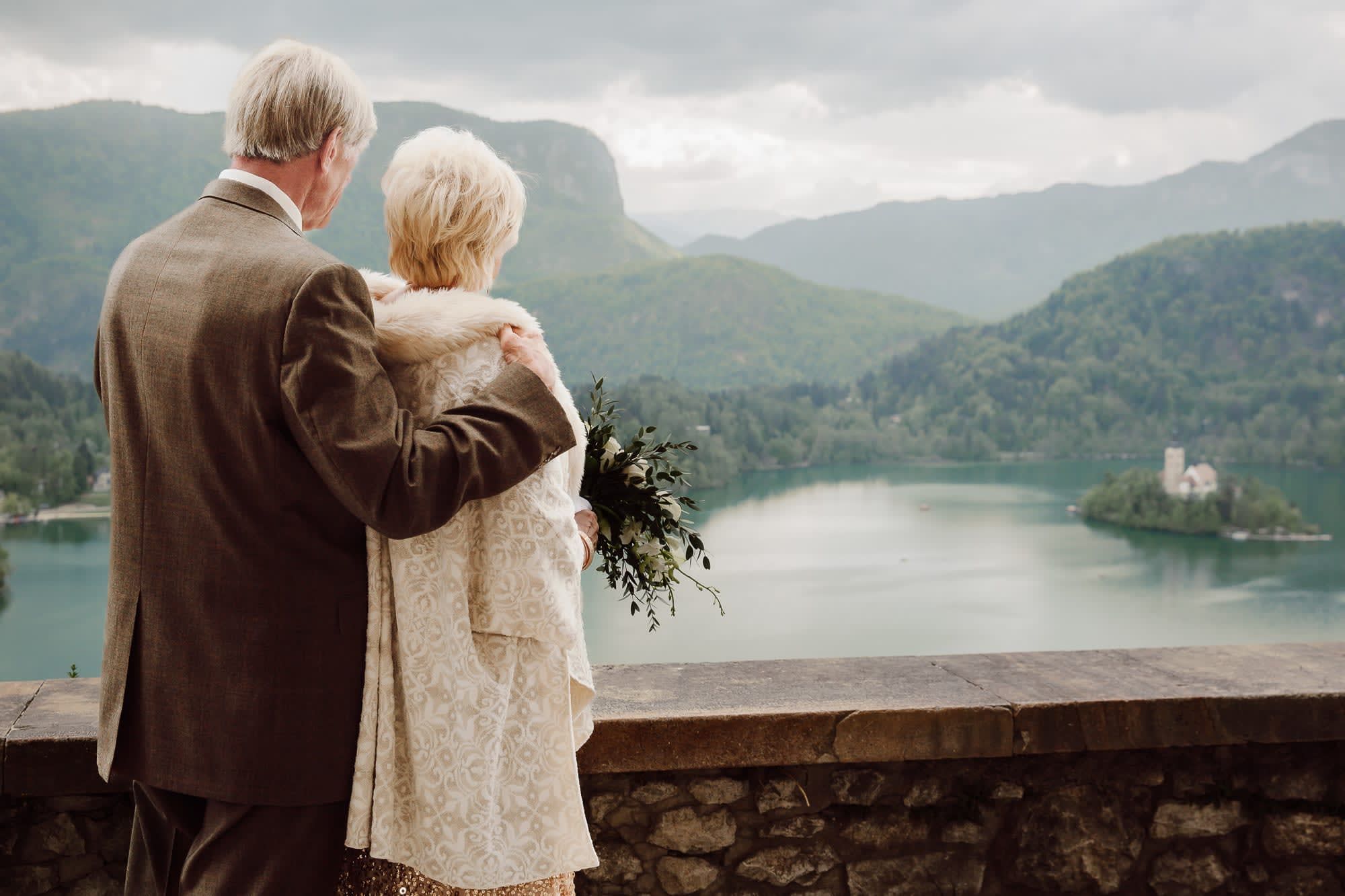 A couple standing together on a quiet terrace overlooking Lake Bled, the island, and the surrounding mountains.
