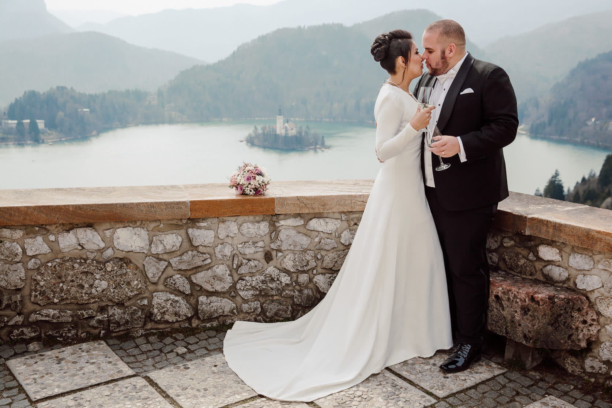 A couple kissing on the terrace of Bled Castle with Lake Bled, Bled Island, and the mountains in the background.