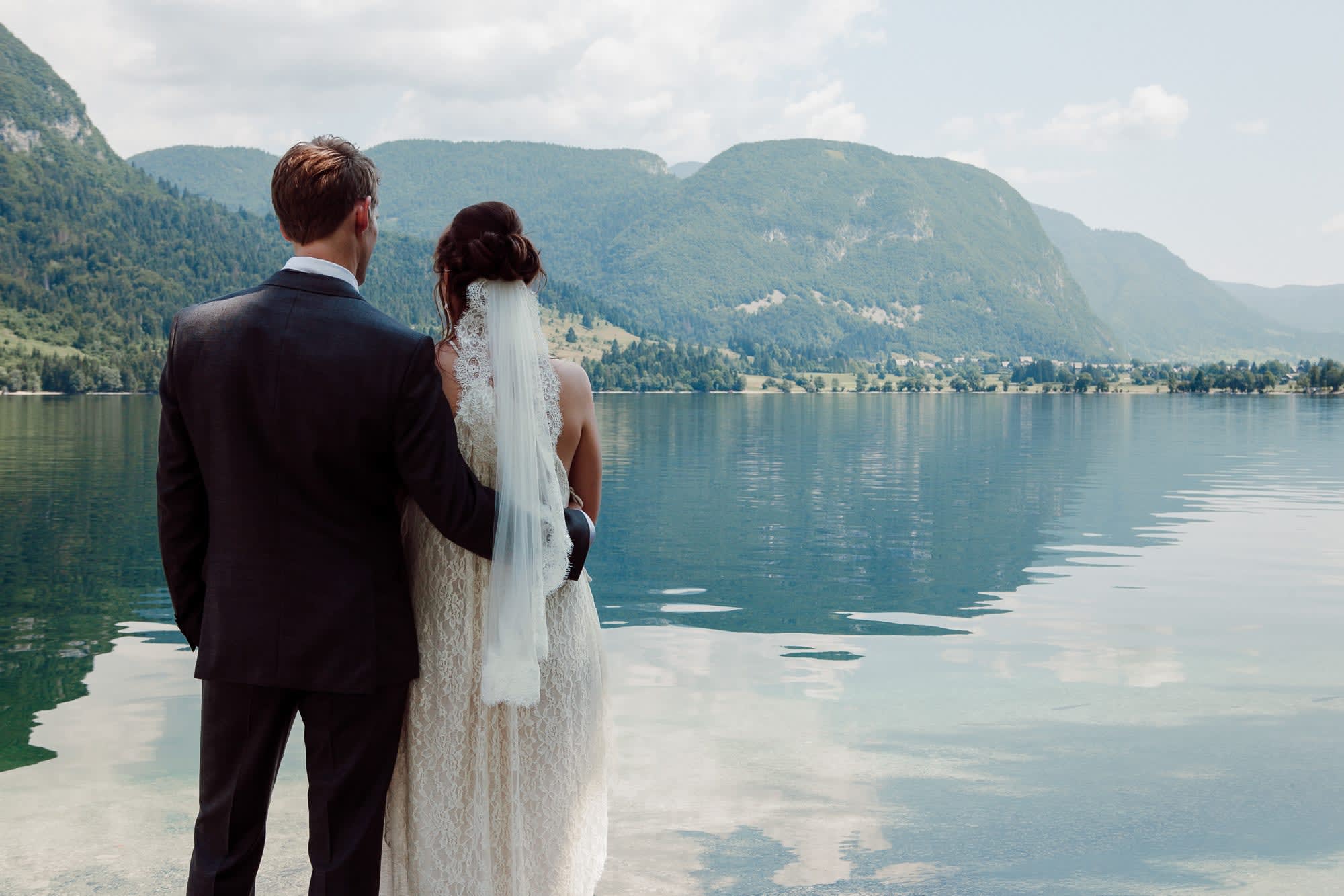 Bride and groom standing together beside a peaceful alpine lake in Slovenia with mountain views across the water.