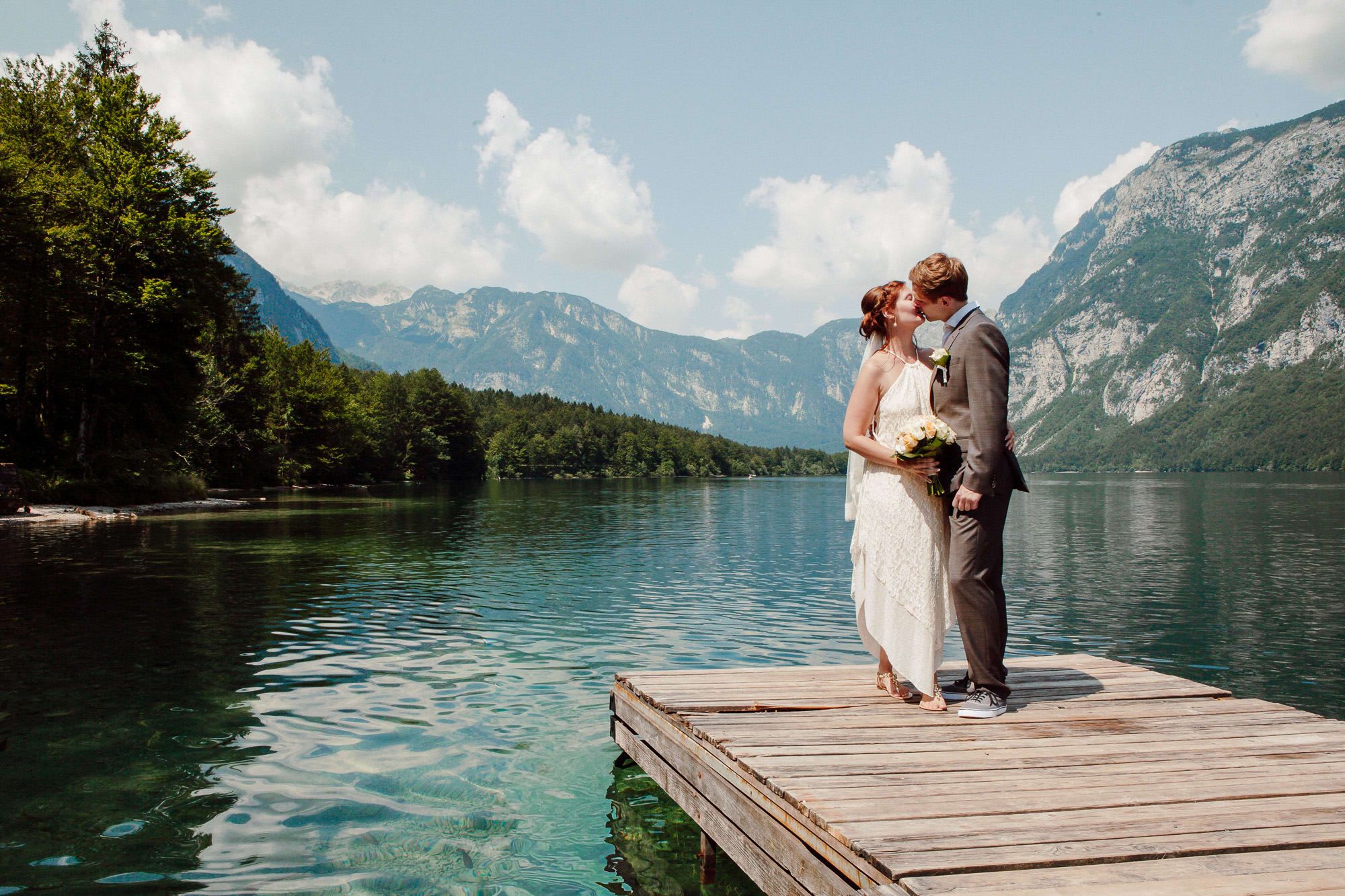 Bride and groom kissing on a wooden dock beside an alpine lake in Slovenia, surrounded by mountains and clear water.