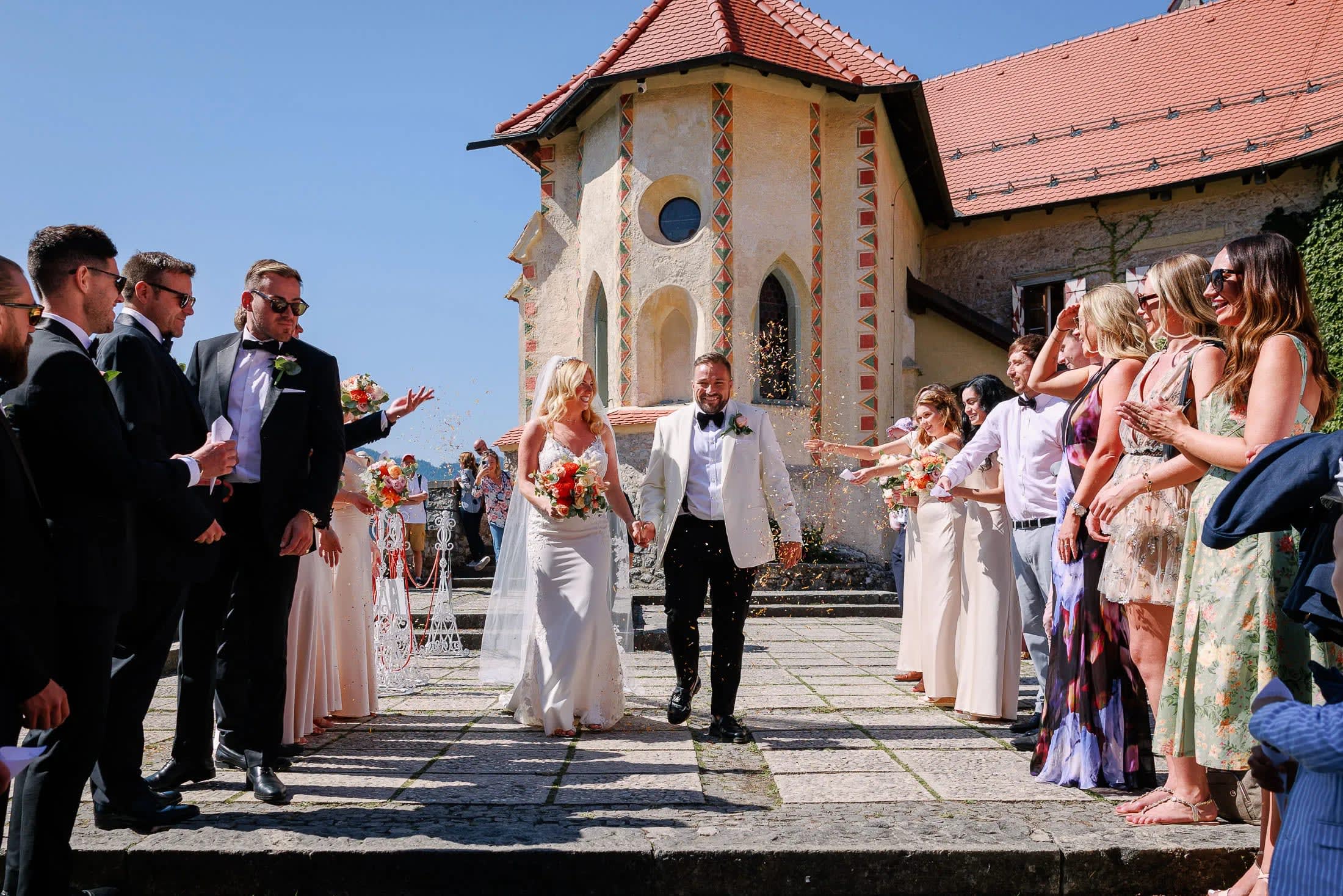 A bride and groom walking hand in hand outside a church, surrounded by joyful guests throwing confetti.
