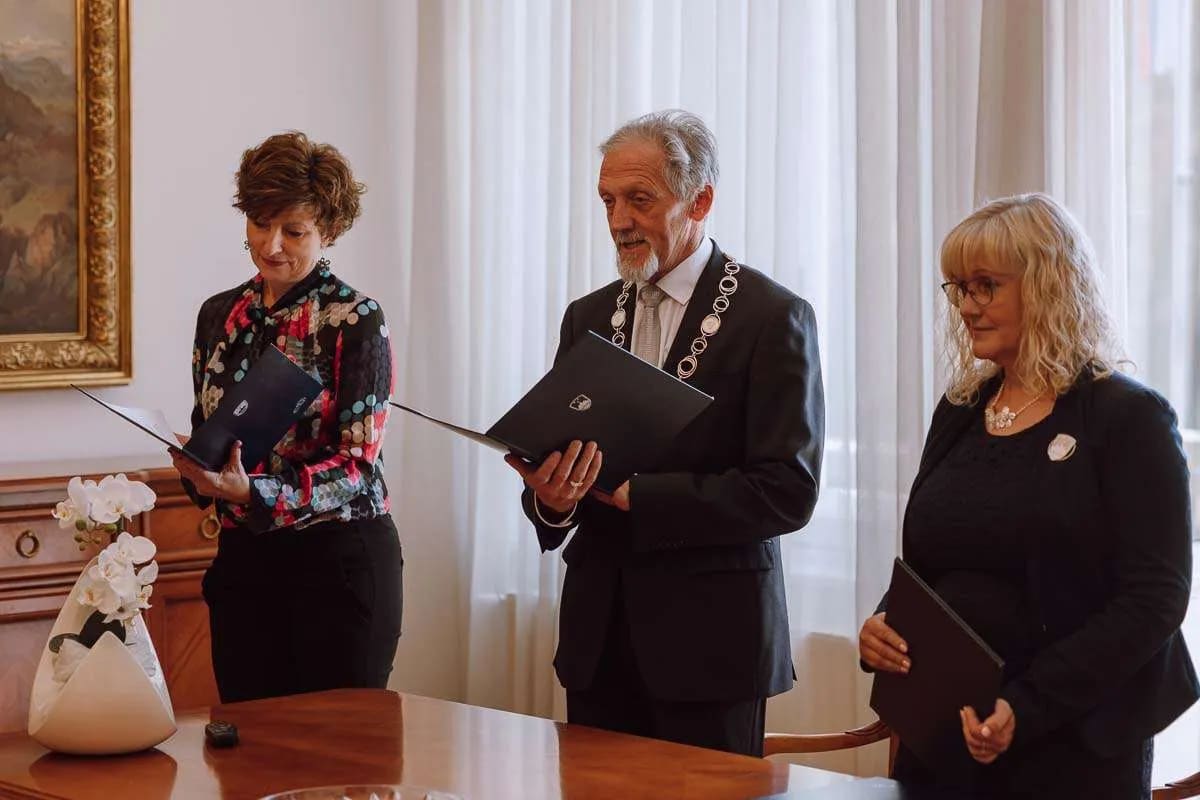 Three individuals standing at a wedding ceremony, holding folders and appearing to conduct official duties, inside a well-lit room.