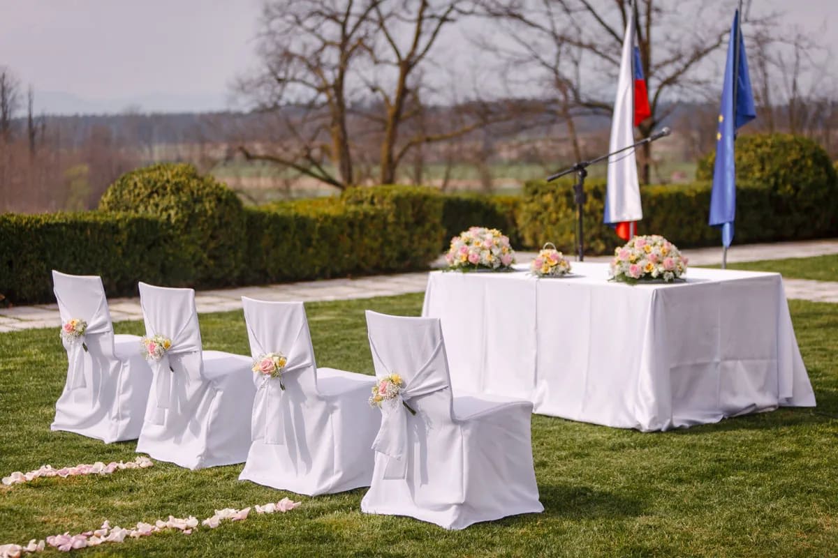 An outdoor wedding ceremony setup with chairs draped in white covers and decorated with flowers, facing a table with floral arrangements and flags in the background.