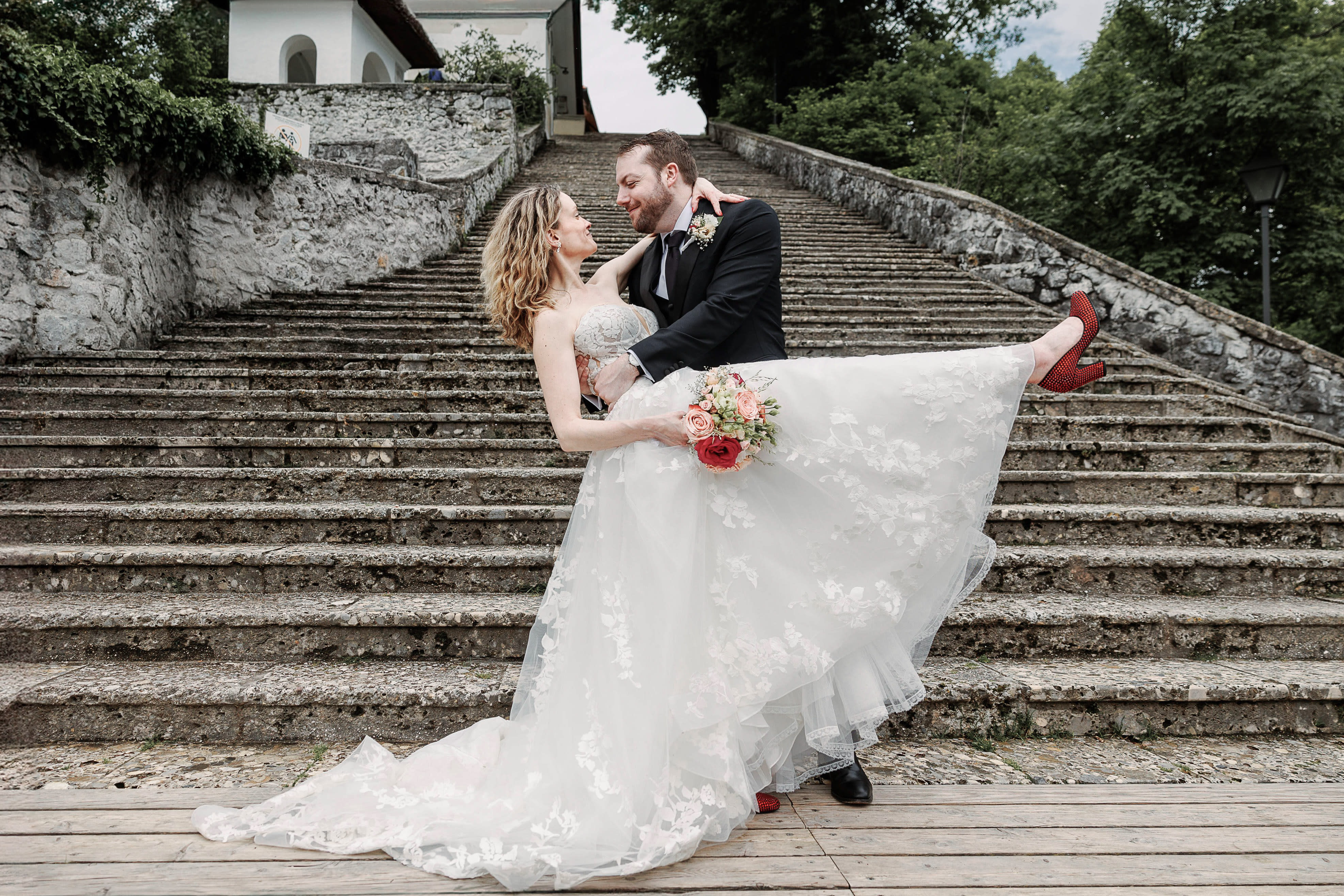 Groom holding bride in front of the 99 steps on Bled Island, Slovenia, both smiling lovingly as she lifts one leg in red heels and holds a bouquet of roses.