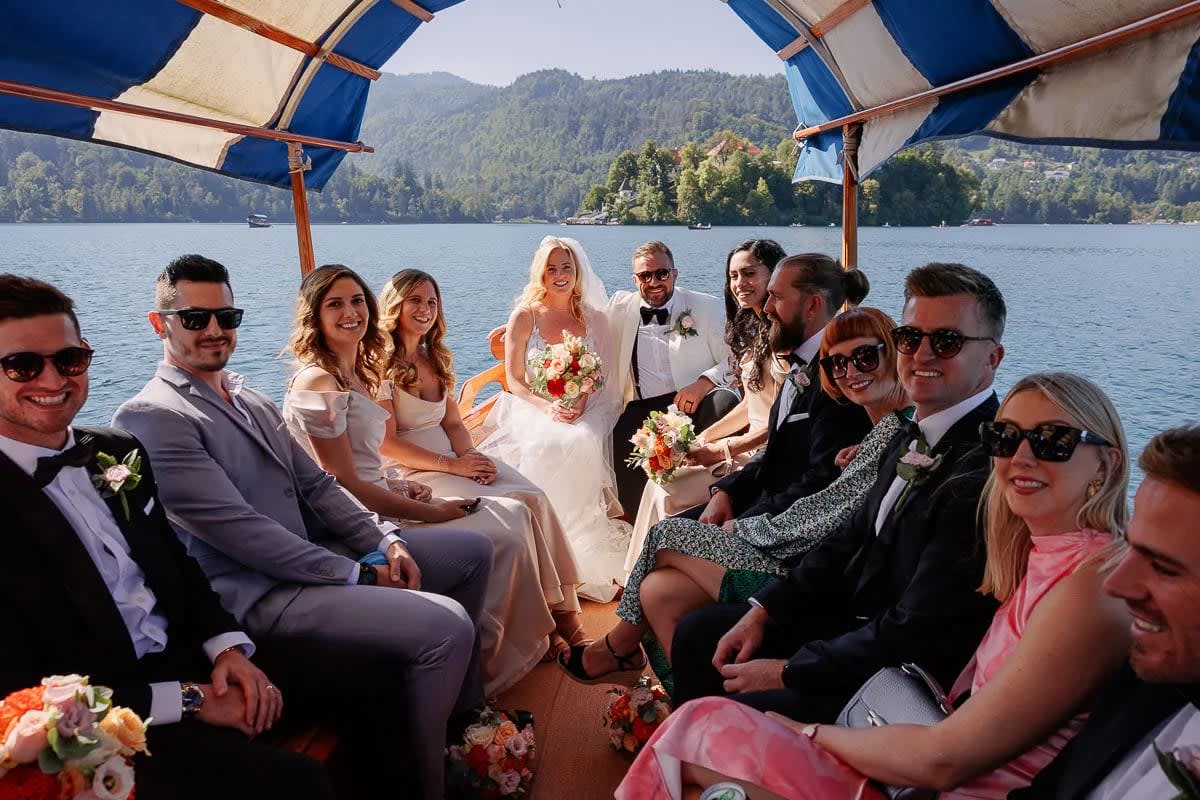 A wedding party, including the bride and groom, seated in a boat on Lake Bled, with mountains in the background.
