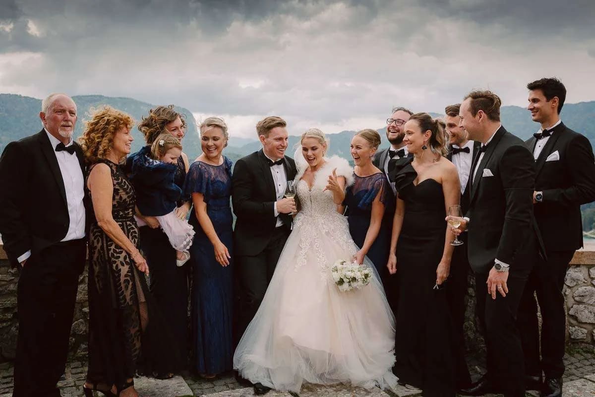 A wedding party with the bride and groom surrounded by family and friends, all dressed in formal attire with a mountain view in the background.