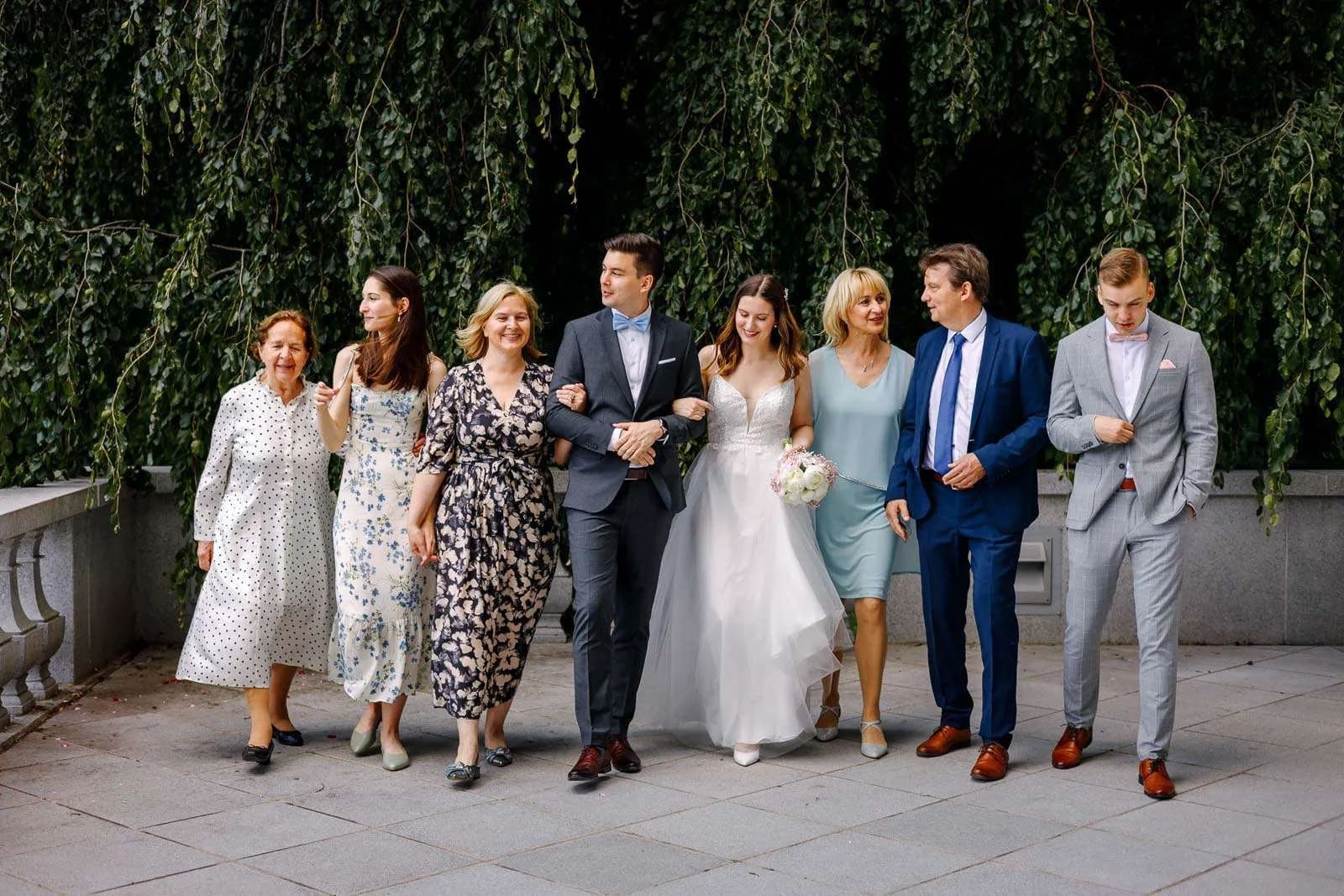  A bride and groom walking arm in arm with family and friends, all smiling and dressed in formal attire, in front of lush greenery.