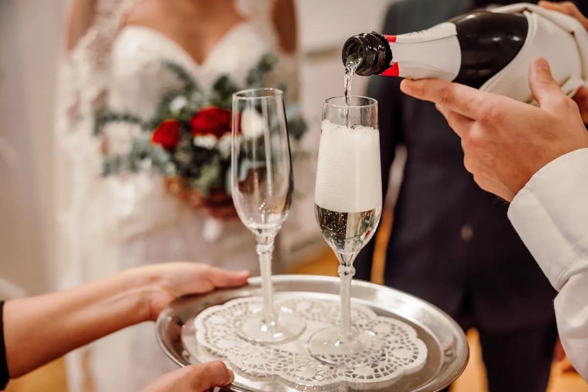 A close-up of champagne being poured into flutes on a silver tray during a wedding celebration, with a bride holding a bouquet in the background.
