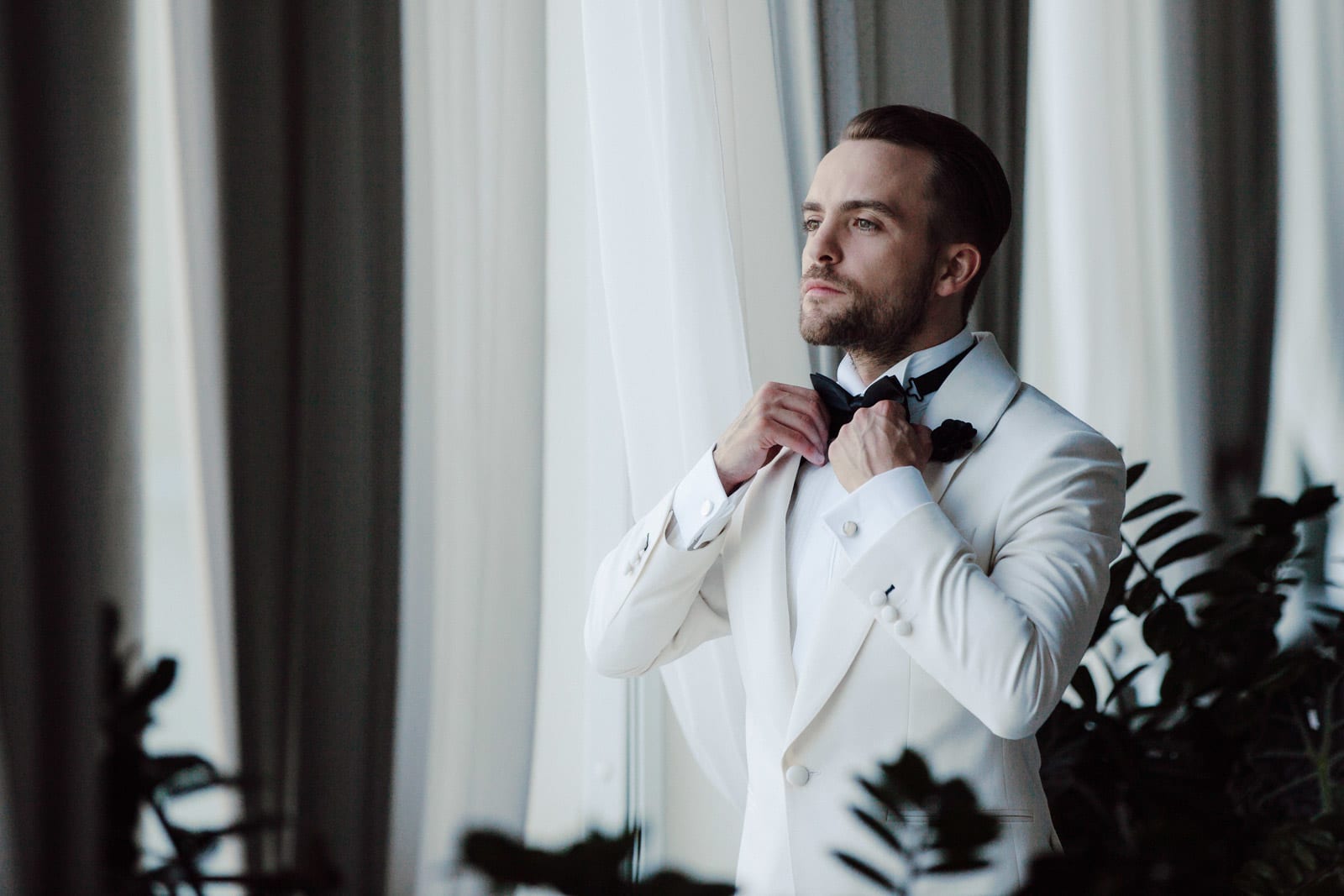 Groom adjusting his tie in soft window light, refined prep portrait.