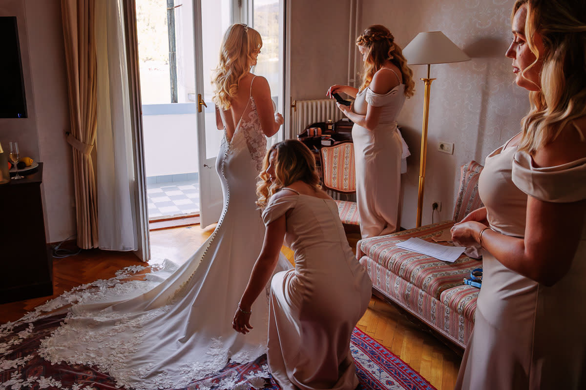 A bride in a lace wedding gown getting ready with the help of bridesmaids in a sunlit room.