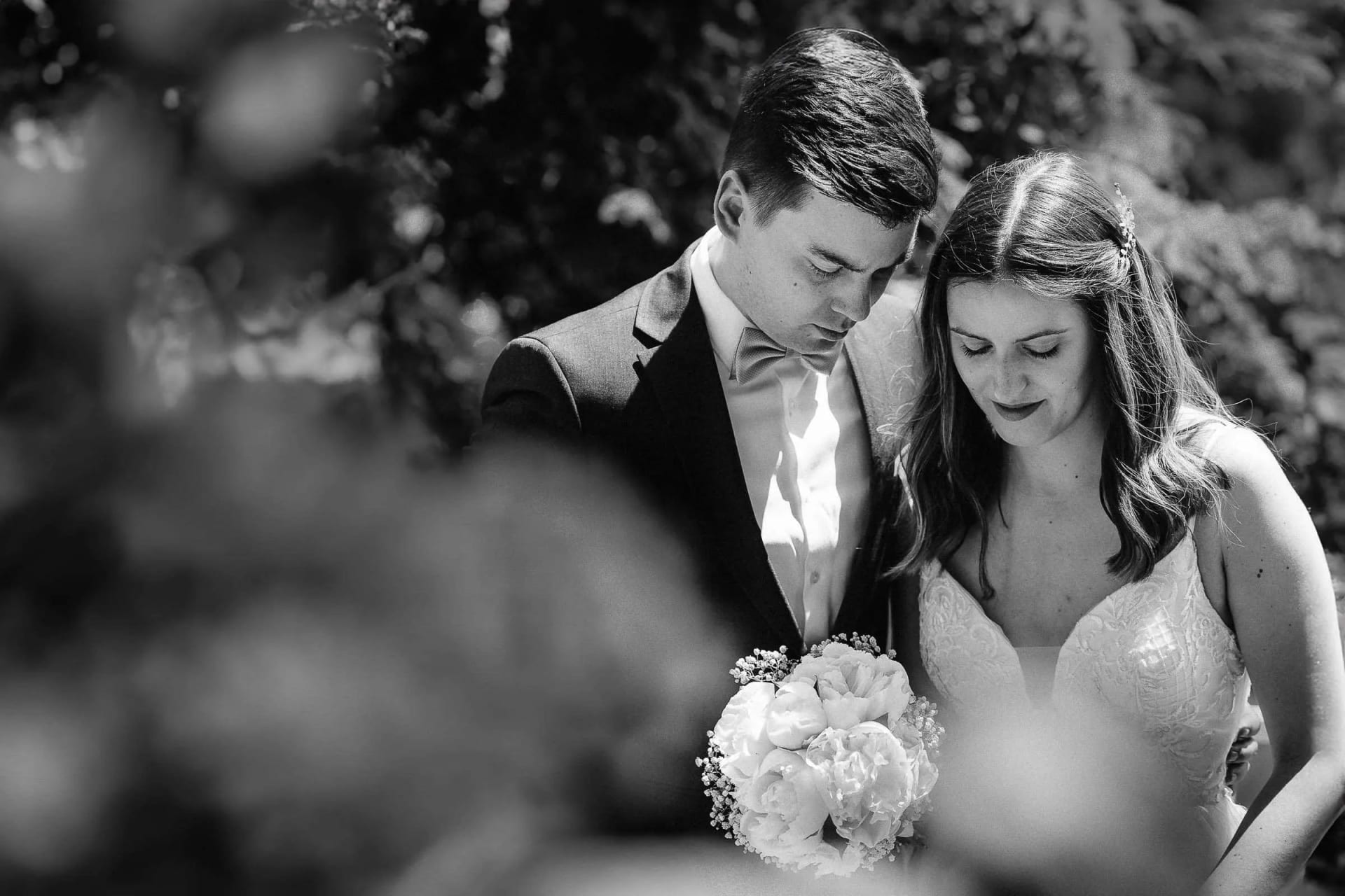 A bride and groom standing closely together, holding a bouquet of flowers, captured in a tender black and white moment.