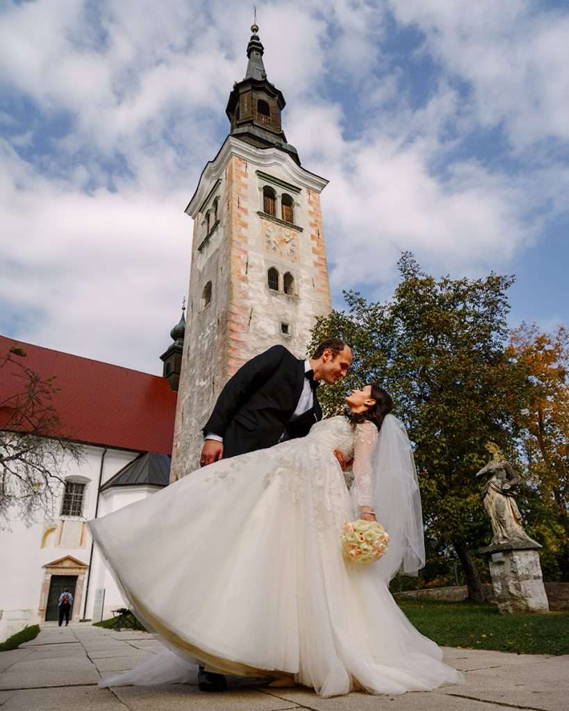 A bride and groom sharing a romantic moment in front of a tall Bled island church tower, with autumn trees surrounding them.