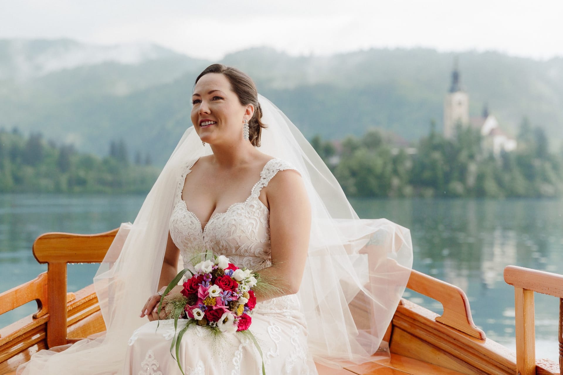 A bride sitting in a wooden pletna boat on Lake Bled with her veil, bouquet, and the island church softly visible in the background.
