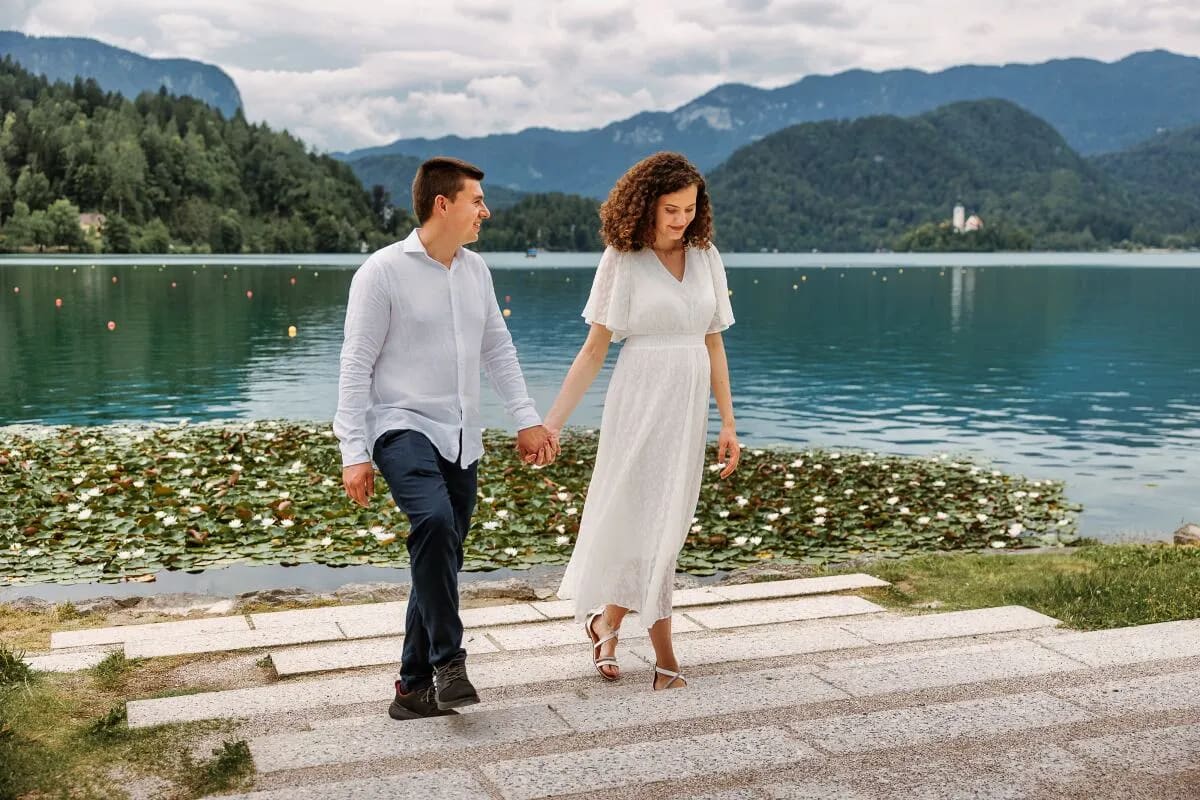 A couple walking hand in hand by the lake with scenic mountains and an island visible in the background.
