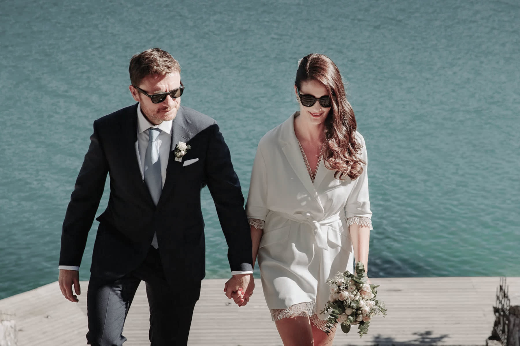 A bride and groom in formal attire holding hands while walking up a dock with a boat in the background on Lake Bled.