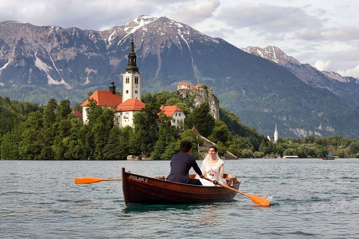 A bride and groom sitting in a rowboat on Lake Bled with the island church and mountains in the background.
