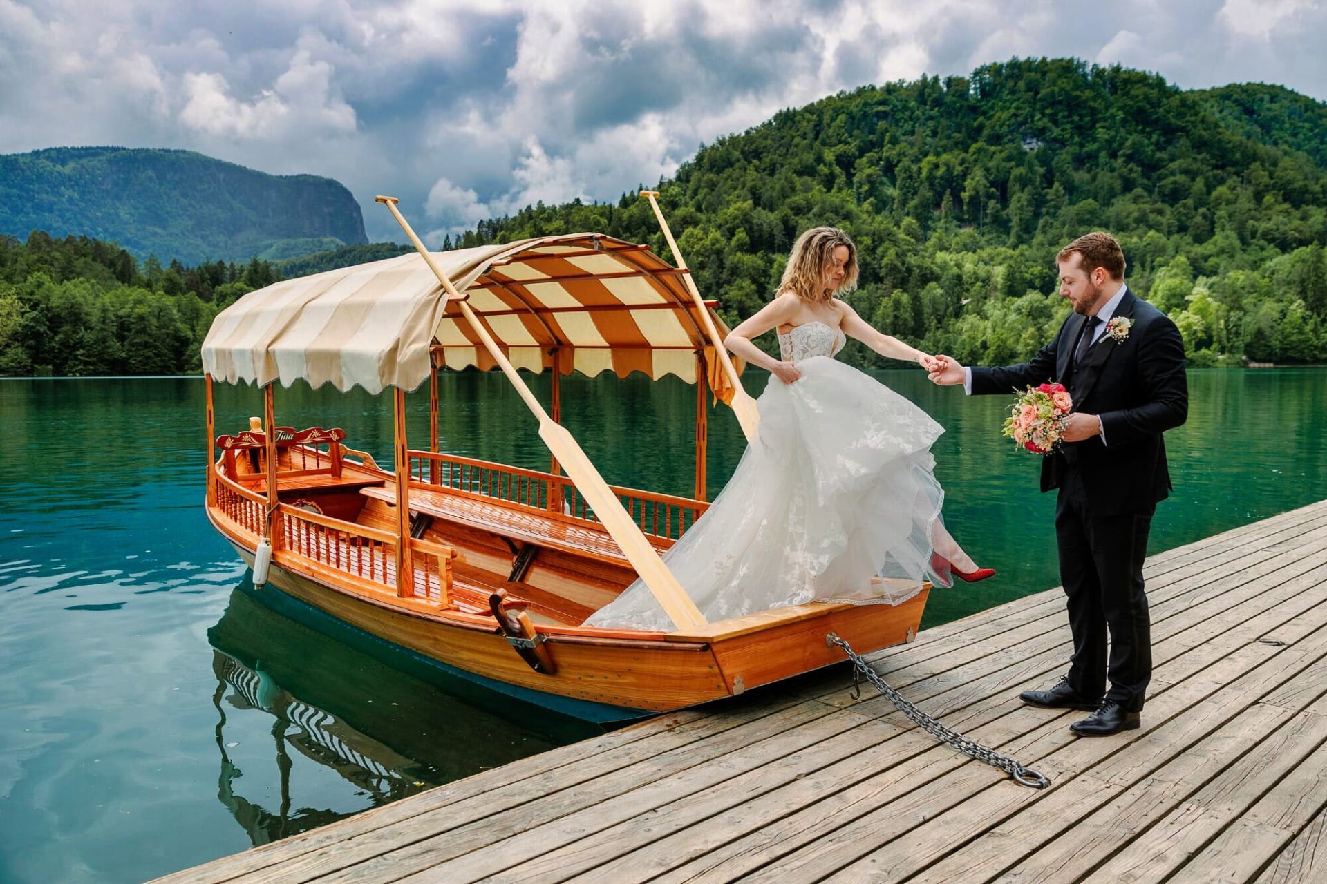 A bride in a white gown steps off a wooden boat onto a dock, assisted by the groom holding a bouquet, with lush green hills and calm water in the background.