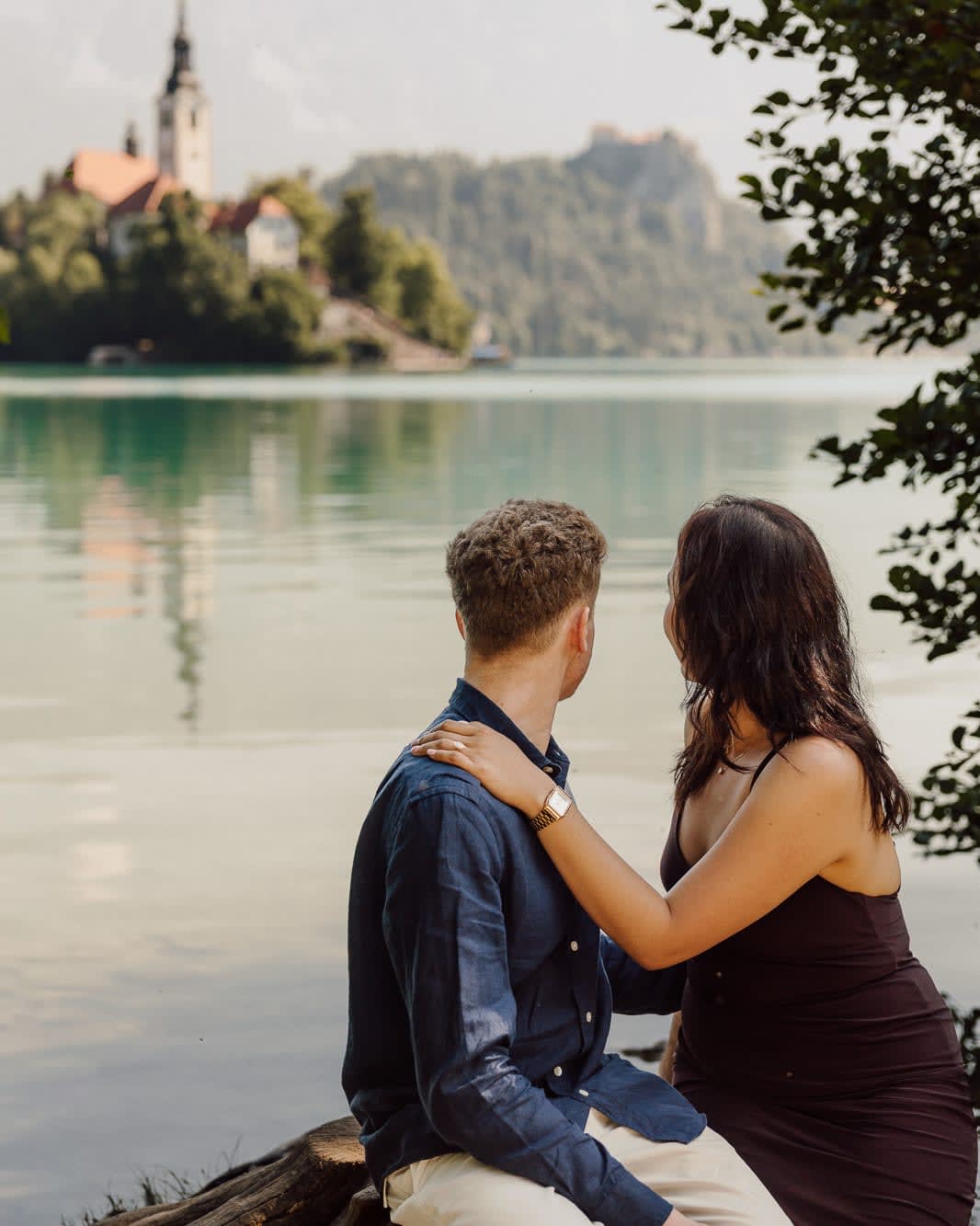 Couple sitting together after the proposal by Lake Bled with Bled Island and Bled Castle in the background.