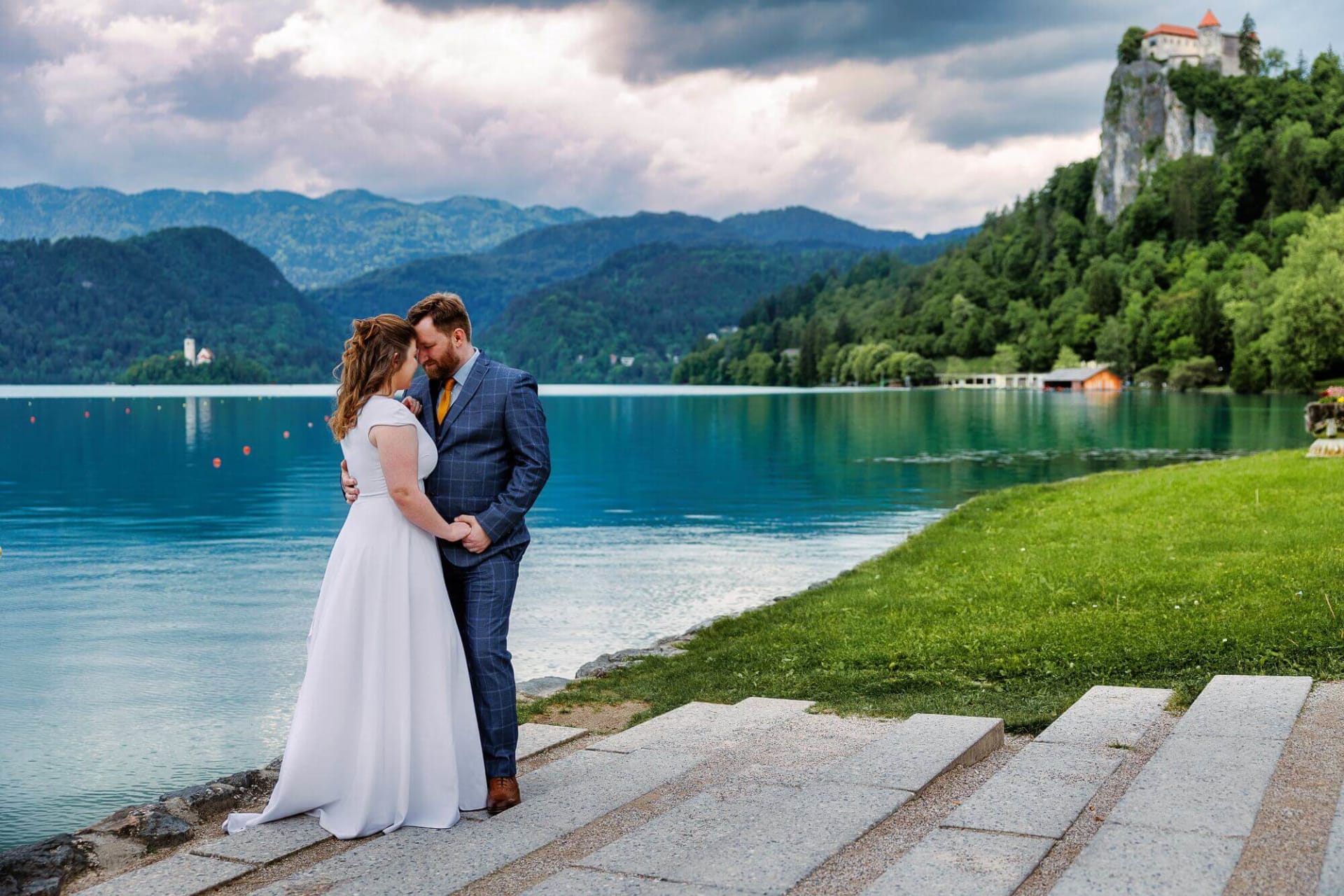 A bride and groom sharing a tender moment by the serene waters of Lake Bled, with a view of the castle and mountains in the background.