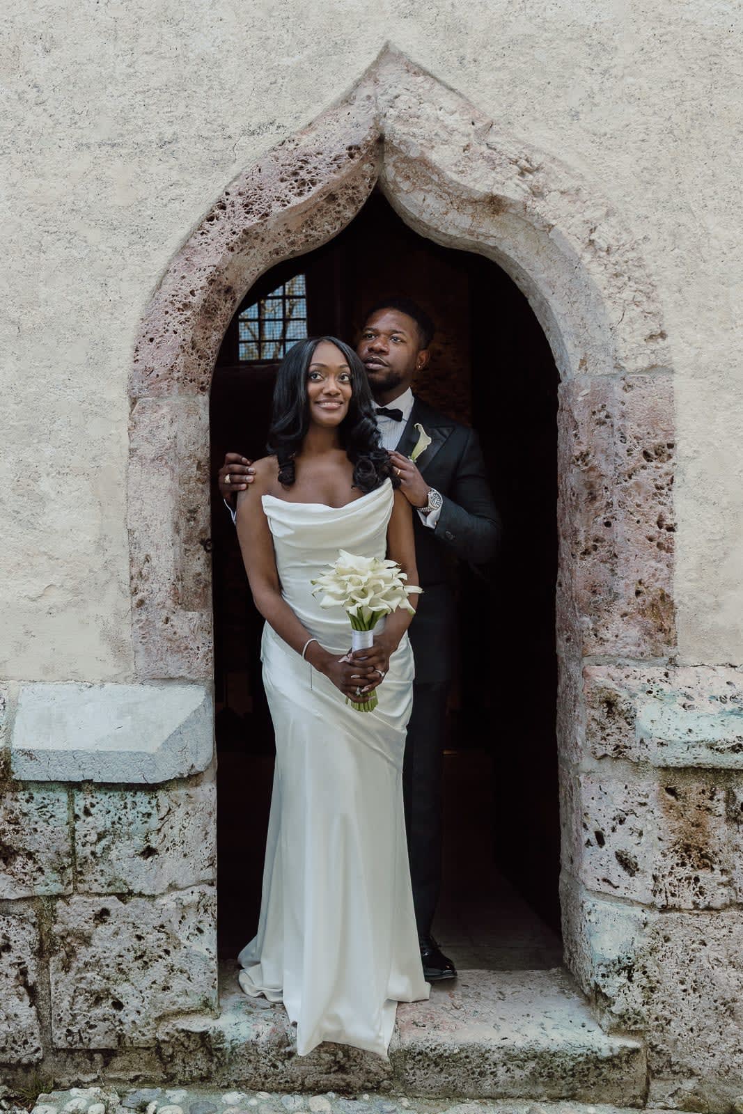 Elopement couple posing in a historic stone doorway at Lake Bled, with the bride holding a white calla lily bouquet.