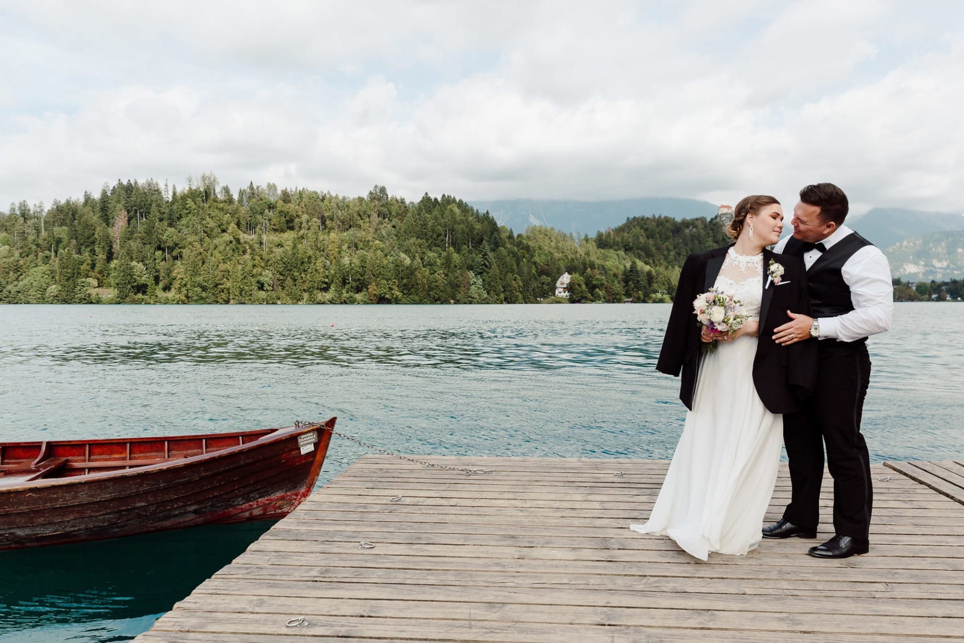 A bride and groom standing together on a wooden dock beside a boat, with Lake Bled and forested hills behind them.