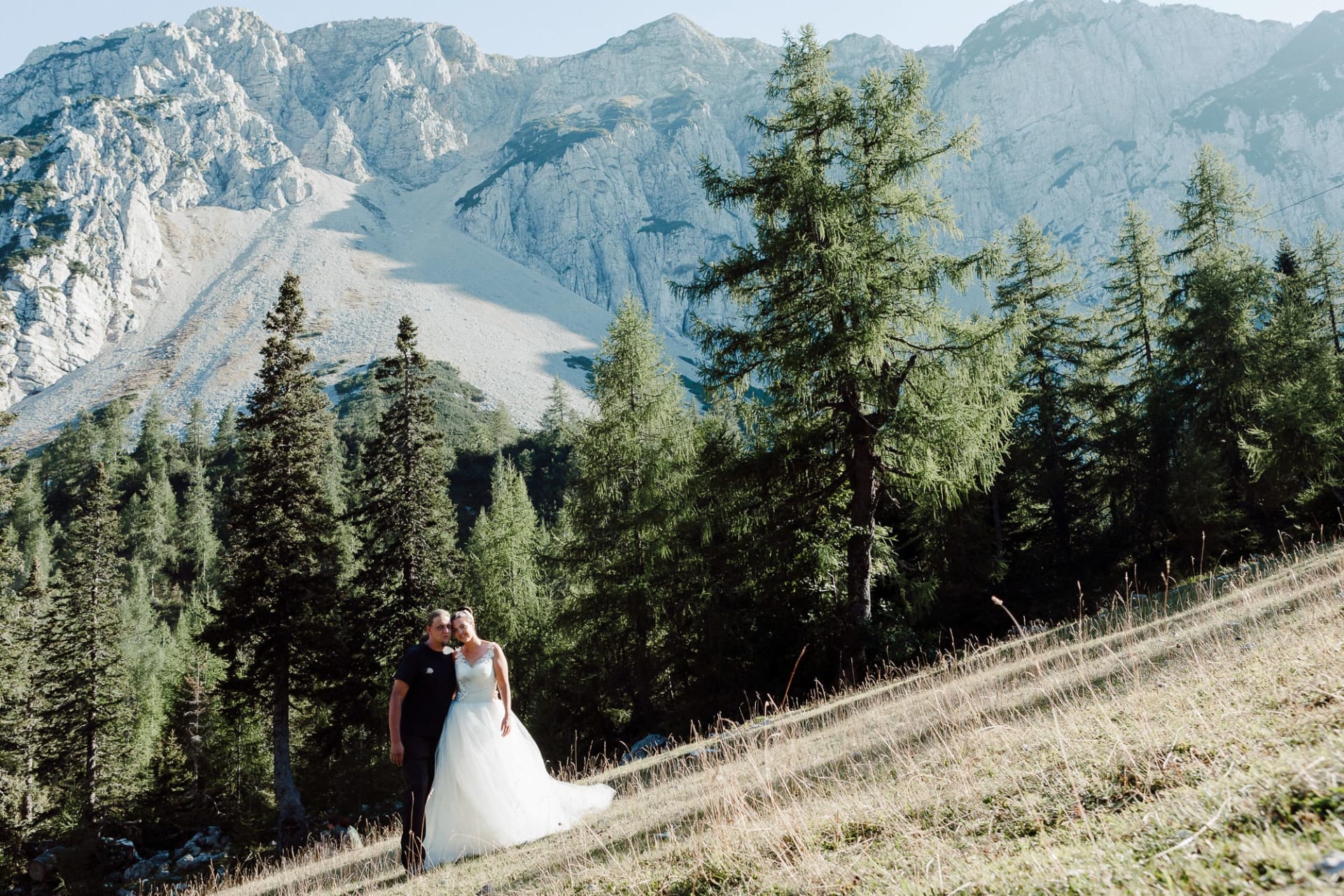 Bride and groom standing together in an alpine meadow in Slovenia with dramatic mountain peaks behind them.