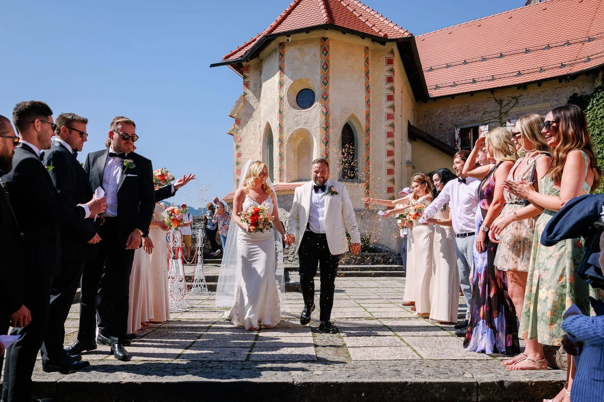 A bride and groom walking hand in hand outside a church, surrounded by joyful guests throwing confetti.