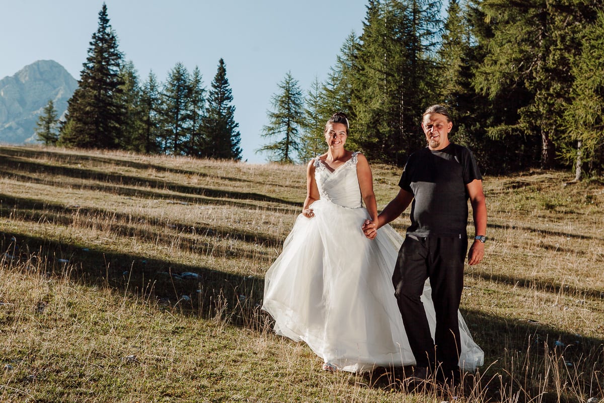 Smiling bride and groom walking hand in hand across an alpine meadow below larches and a mountain peak at Zelenica in the Karavanke, Slovenia.
