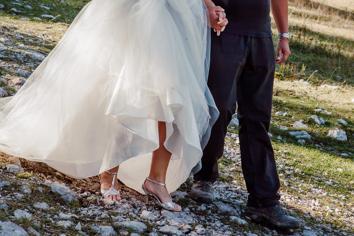 Bride in silver heels and groom in hiking boots walking hand in hand over rocky ground at Zelenica in the Karavanke, Slovenia