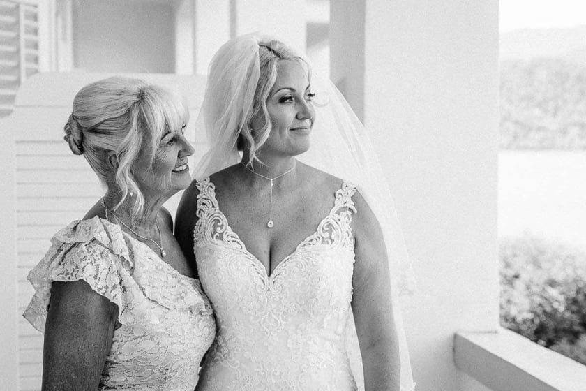 A bride in a lace wedding dress stands next to her mother, both smiling and looking into the distance indoors.