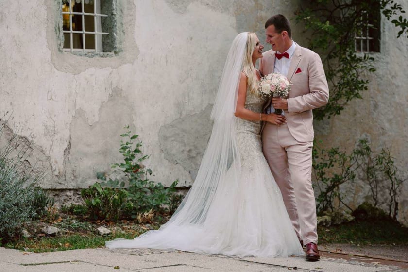 A bride and groom smiling at each other while standing in front of an old stone wall, with the bride holding a bouquet of flowers.