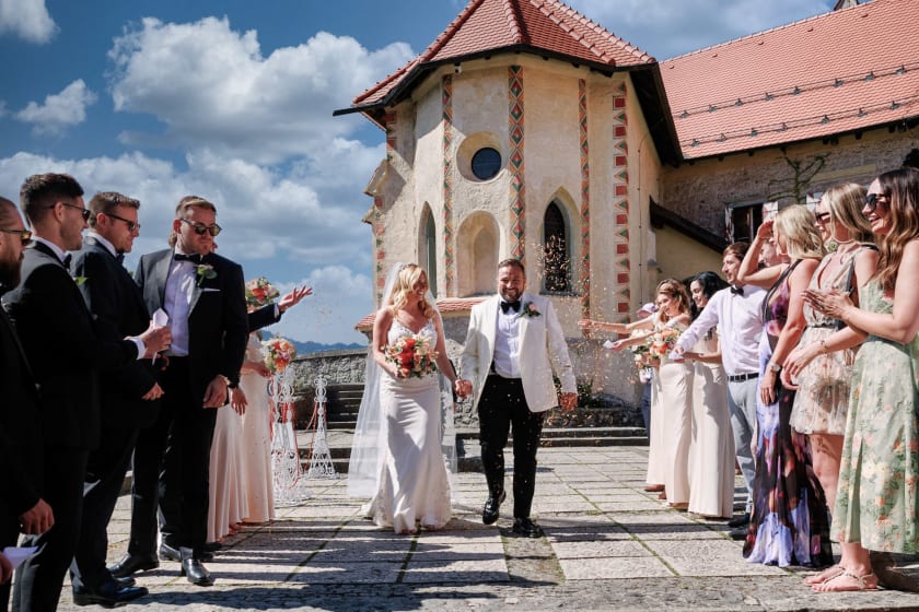 Bride and groom walk hand in hand through a cheering crowd of guests throwing confetti at their wedding ceremony outside Bled Castle. The groom wears a white tuxedo jacket and the bride holds a bouquet while smiling, surrounded by joyful friends and family in elegant attire under a bright blue sky.