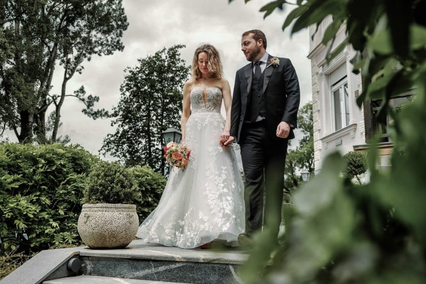 A bride and groom holding hands and walking down steps surrounded by lush greenery and flowers.