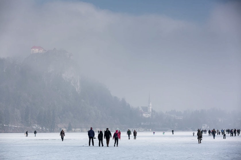 People walking on a frozen Lake Bled with a view of the castle perched on a hill and a church in the background on a misty day.
