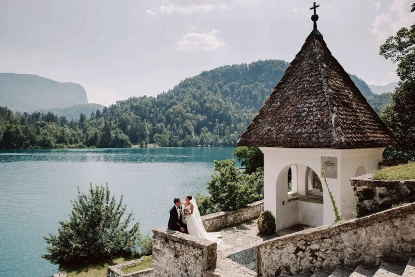 Bride and groom sit on stone steps on Bled island beside a small chapel overlooking Lake Bled, surrounded by lush green hills on a sunny summer day.