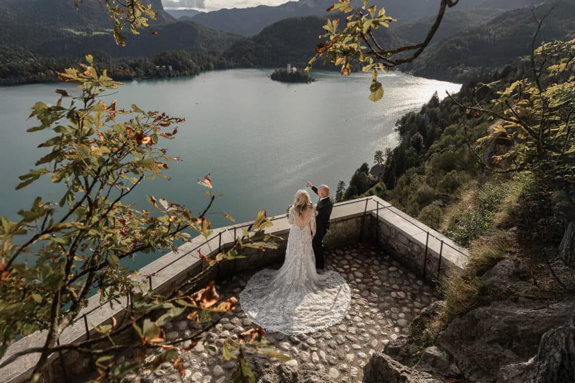 A bride and groom stand on a stone balcony overlooking the scenic Lake Bled with a small island and church in the distance.
