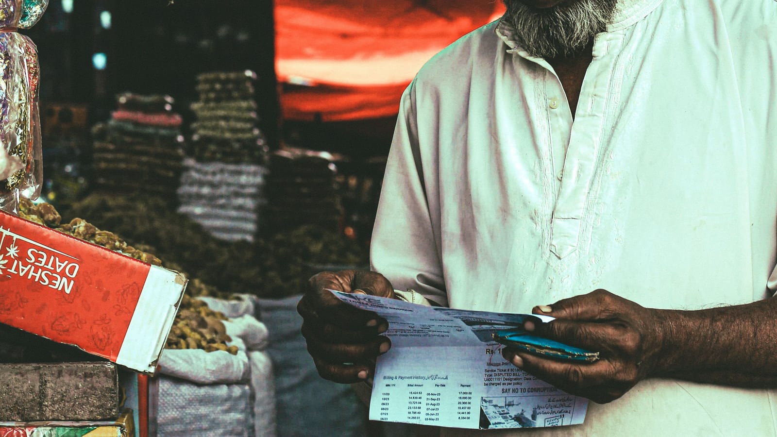 A trader checking a paper list at a dry fruit market, with stock in sacks beside him