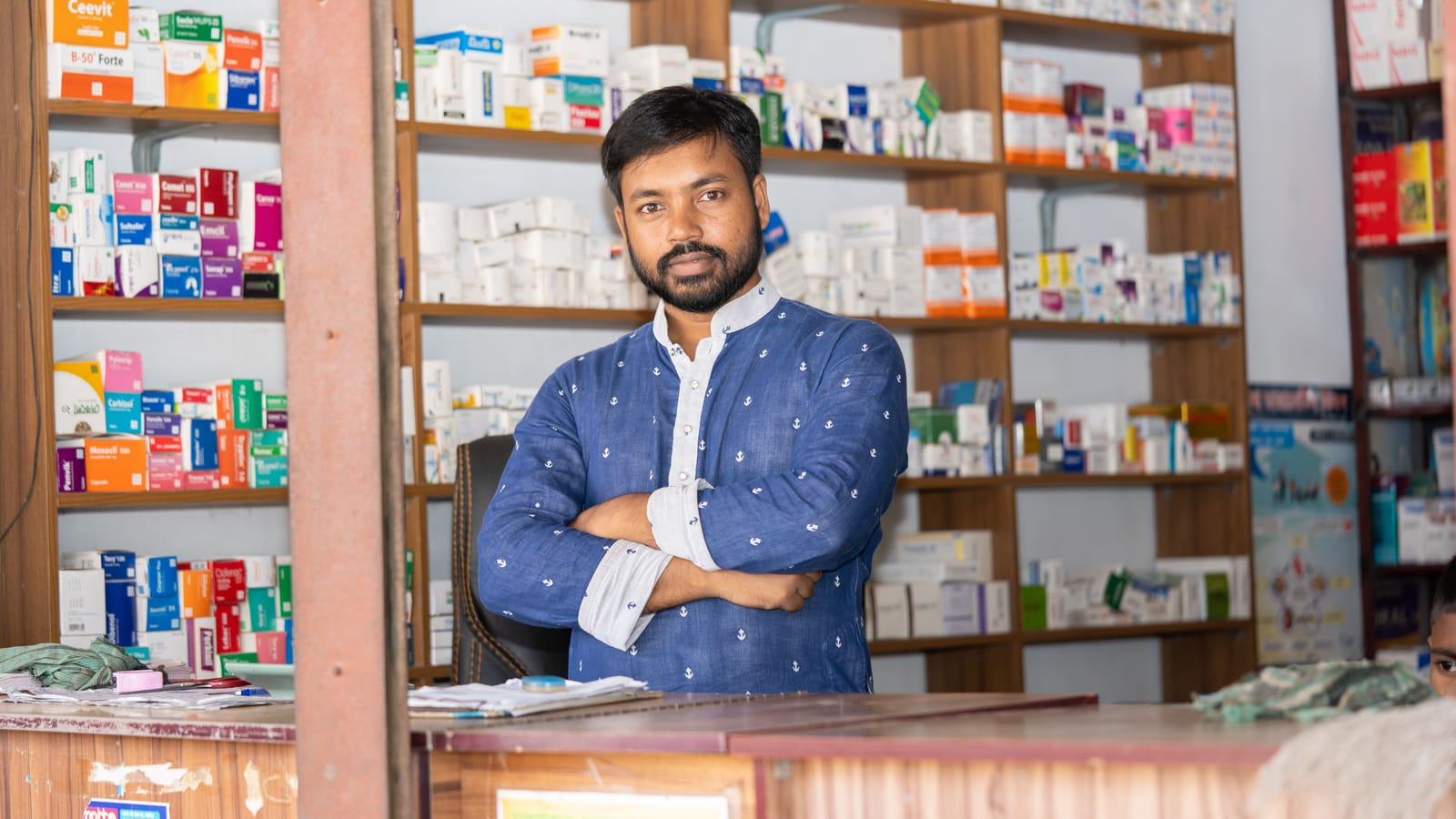 A pharmacist standing behind the counter of a medical store with medicine shelves behind him