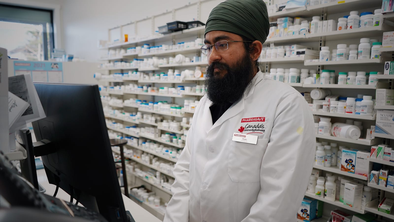 A pharmacist at the computer behind a pharmacy counter, with medicine shelves behind him