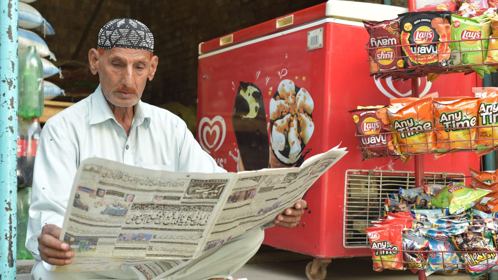 A shopkeeper in Multan reading a newspaper at his small store, with snacks and a freezer beside him