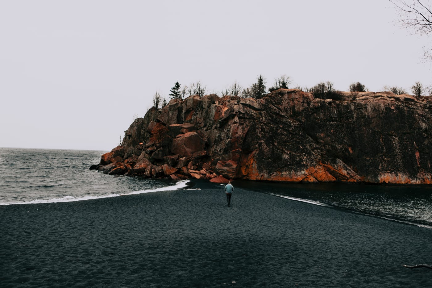 A tiny figure on the beach beneath a towering red rock cliff on the North Shore