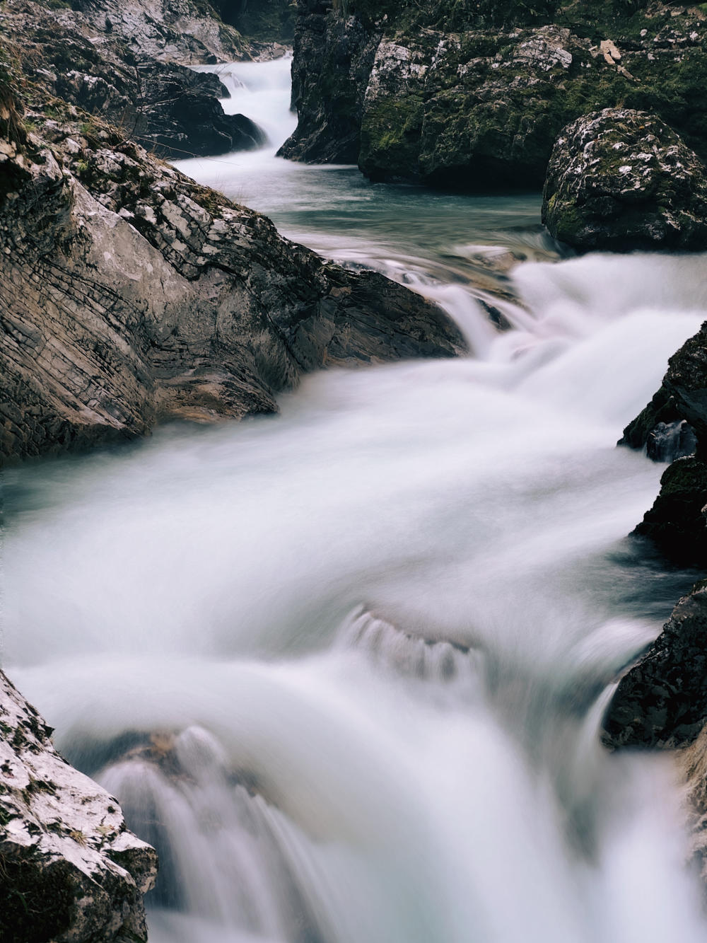 A long exposure of white water running through a gorge. The water has blended into a creamy appearance. The rocks on either side are grey and brown, and are some are covered in moss. Some rocks are polished smooth by the erosion of the water, while others show striation lines and scars from years of movement.