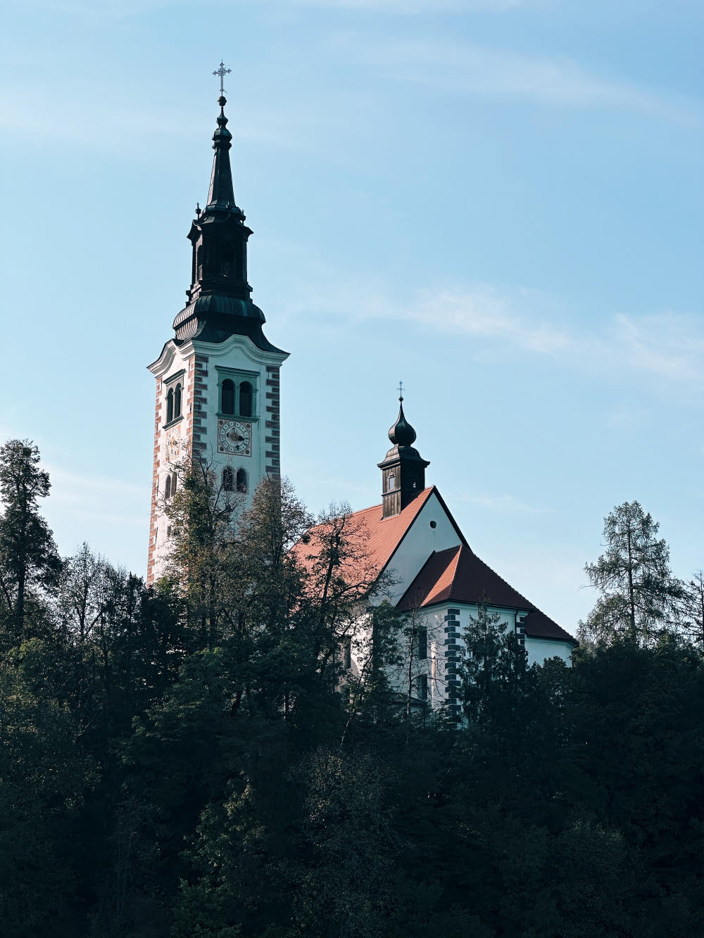 The spire of the Church of the Mother of God on the Lake, in Bled, Slovenia. The pitched roof features orange tiles, above white plaster, and reddish brick work. The spire is ornate and made of metal. Trees run around the circumference of the island and feature in the foreground of the photograph.