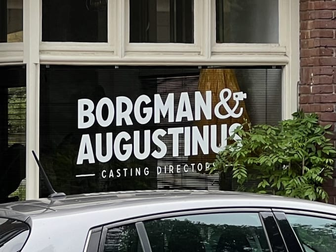A office window signpainted with the phrase 'Borgman & Augustinus Casting Directors'. The Ampersand is holding a camera.