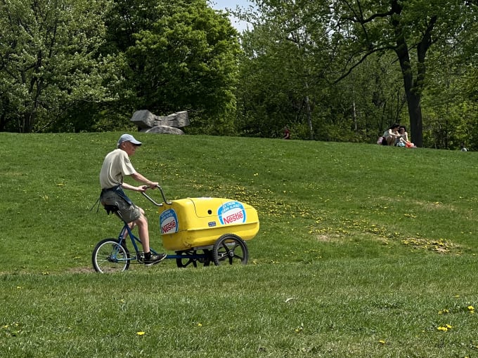 An ice cream man riding his ice cream bike through a park