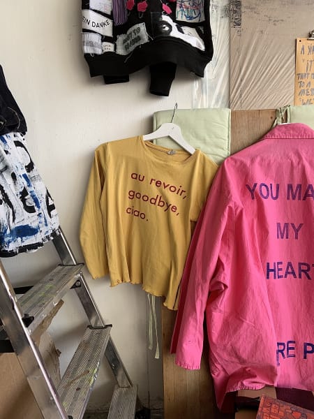 A yellow cotton three quarter sleeve top hanging on a wall with the text au revoir, goodbye, ciao printed on it in red