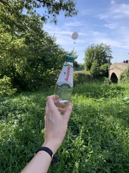 Photo me holding up a small glass water bottle in front of a green space with an air balloon in background