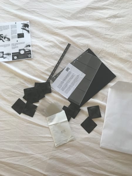 Coin collecting supplies including black mounting pieces, clear mini vinyl slips spilling from a small white envelope printed with '812', a gridded vinyl binder sleeve to hold the mounts and individual slips, a black binder separator, instructions and the large white envelope it arrived in strewn on a powder pink bedsheet.